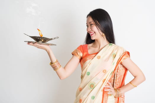 Portrait of young mixed race Indian Chinese female in traditional sari dress, holding diya oil lamp light, on plain background.