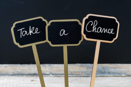Motivational message Take A Chance written with chalk on wooden mini blackboard labels, defocused chalkboard and wood table in background