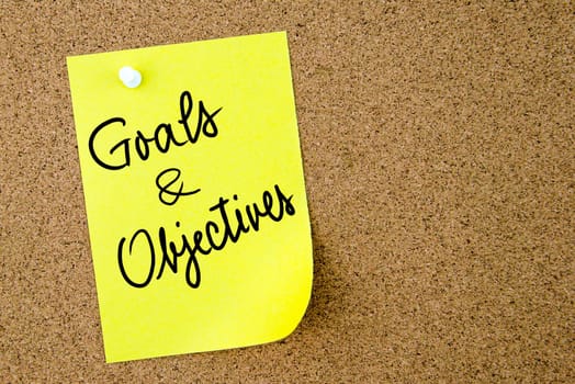 Goals and Objectives text written on yellow paper note pinned on cork board with white thumbtack. Business concept image with copy space available