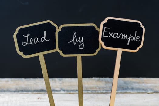 Business message Lead By Example written with chalk on wooden mini blackboard labels, defocused chalkboard and wood table in background