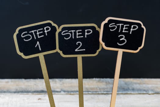 Business message Step 1, Step 2, Step 3 written with chalk on wooden mini blackboard labels, defocused chalkboard and wood table in background