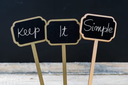Motivational message Keep It Simple written with chalk on wooden mini blackboard labels, defocused chalkboard and wood table in background