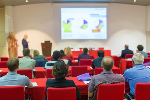 Speaker Giving a Talk at Business Meeting. Audience in the conference hall. Business and Entrepreneurship. Focus on unrecognizable people from rear.