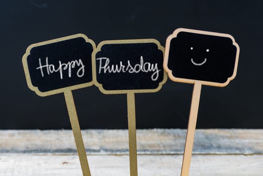 Motivational message Happy Thursday written with chalk on wooden mini blackboard labels, defocused chalkboard and wood table in background