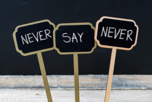 Motivational message NEVER SAY NEVER written with chalk on wooden mini blackboard labels, defocused chalkboard and wood table in background