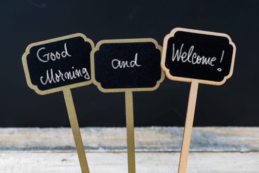 Greeting message Good Morning and Welcome written with chalk on wooden mini blackboard labels, defocused chalkboard and wood table in background