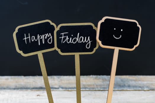 Motivational message Happy Friday written with chalk on wooden mini blackboard labels, defocused chalkboard and wood table in background