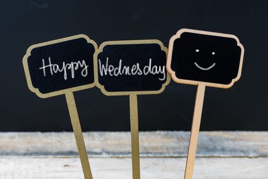Motivational message Happy Wednesday written with chalk on wooden mini blackboard labels, defocused chalkboard and wood table in background