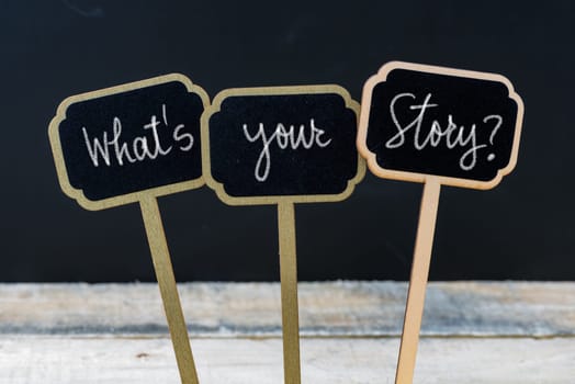 Motivational message Whats Your Story ? written with chalk on wooden mini blackboard labels, defocused chalkboard and wood table in background