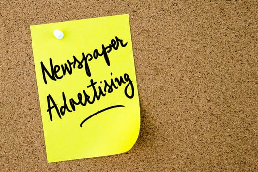 Newspaper Advertising text written on yellow paper note pinned on cork board with white thumbtack. Business concept image with copy space available