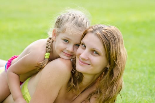 Daughter lying on mother's back happily hugs her, against the background of green grass