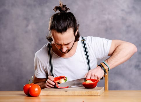 young bearded man cutting sweet pepper in front of gray background