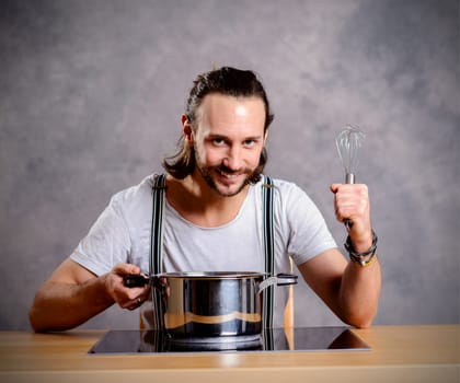 young bearded man with cooking pot and egg whish in front of gray background