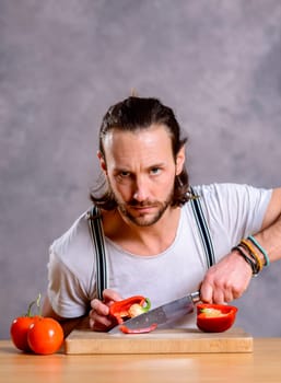 young bearded man cutting sweet pepper in front of gray background