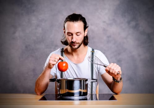 young bearded man with cooking pot, eggwhish and tomato in front of gray background