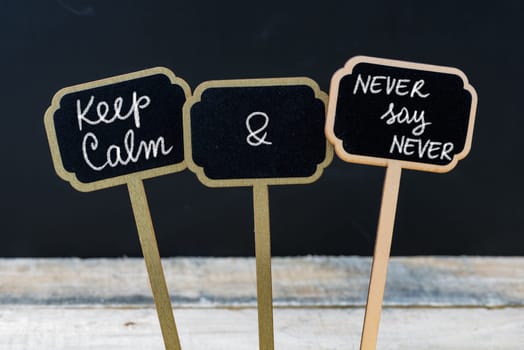 Keep Calm and Never Say Never message written with chalk on mini blackboard labels, defocused chalkboard and wooden table in background. Fun and humor concept