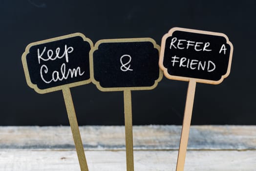 Keep Calm and Refer A Friend message written with chalk on mini blackboard labels, defocused chalkboard and wooden table in background. Fun and humor concept