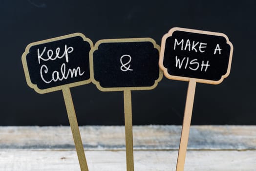 Keep Calm and Make A Wish message written with chalk on mini blackboard labels, defocused chalkboard and wooden table in background. Fun and humor concept