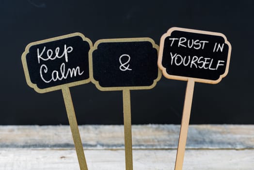 Keep Calm and Trust In Yourself message written with chalk on mini blackboard labels, defocused chalkboard and wooden table in background. Fun and humor concept