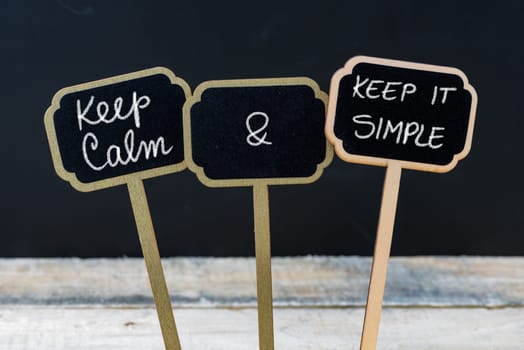 Keep Calm and Keep It Simple message written with chalk on mini blackboard labels, defocused chalkboard and wooden table in background. Fun and humor concept