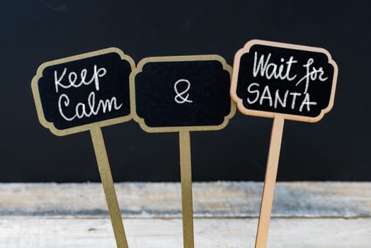 Keep Calm and Wait For Santa message written with chalk on mini blackboard labels, defocused chalkboard and wooden table in background. Fun and humor concept