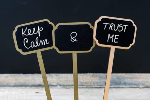Keep Calm and Trust Me message written with chalk on mini blackboard labels, defocused chalkboard and wooden table in background. Fun and humor concept