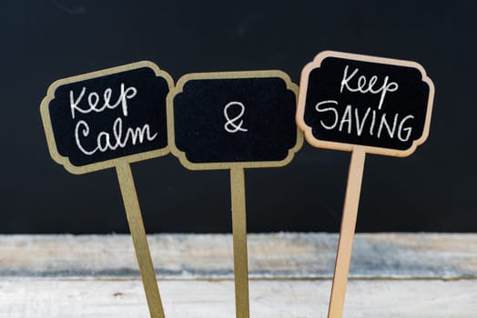 Keep Calm and Keep Saving message written with chalk on mini blackboard labels, defocused chalkboard and wooden table in background. Fun and humor concept