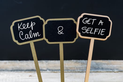 Keep Calm and Get A Selfie message written with chalk on mini blackboard labels, defocused chalkboard and wooden table in background. Fun and humor concept
