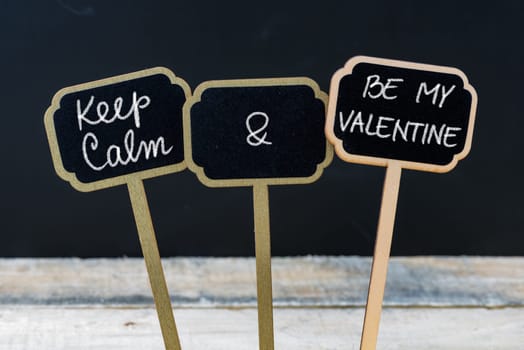 Keep Calm and Be My Valentine message written with chalk on mini blackboard labels, defocused chalkboard and wooden table in background. Fun and humor concept