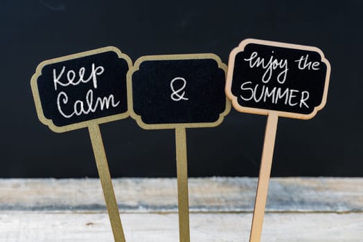 Keep Calm and Enjoy The Summer message written with chalk on mini blackboard labels, defocused chalkboard and wooden table in background. Fun and humor concept