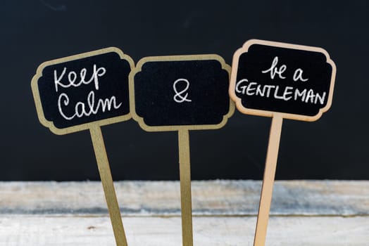 Keep Calm and Be A Gentleman message written with chalk on mini blackboard labels, defocused chalkboard and wooden table in background. Fun and humor concept