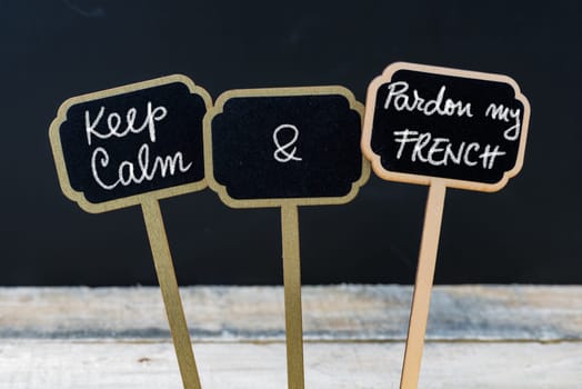 Keep Calm and Pardon My French message written with chalk on mini blackboard labels, defocused chalkboard and wooden table in background. Fun and humor concept