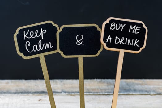 Keep Calm and Buy Me A Drink message written with chalk on mini blackboard labels, defocused chalkboard and wooden table in background. Fun and humor concept