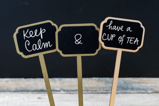 Keep Calm and Have A Cup Of Tea message written with chalk on mini blackboard labels, defocused chalkboard and wooden table in background. Fun and humor concept
