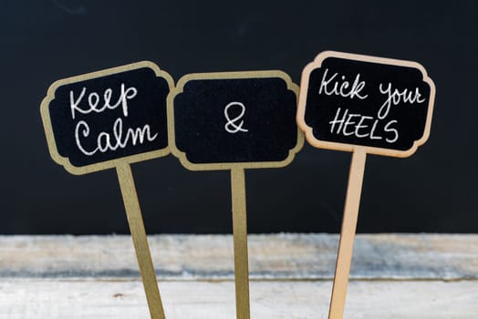 Keep Calm and Kick Your Heels message written with chalk on mini blackboard labels, defocused chalkboard and wooden table in background. Fun and humor concept