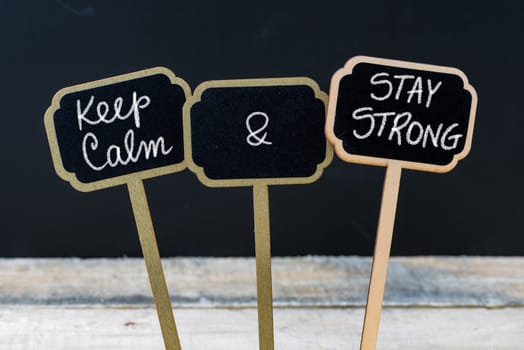 Keep Calm and Stay Strong message written with chalk on mini blackboard labels, defocused chalkboard and wooden table in background. Fun and humor concept