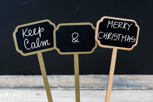Keep Calm and Merry Christmas message written with chalk on mini blackboard labels, defocused chalkboard and wooden table in background. Fun and humor concept