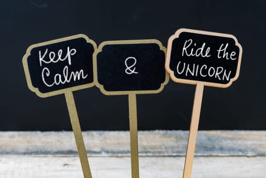Keep Calm and Ride The Unicorn message written with chalk on mini blackboard labels, defocused chalkboard and wooden table in background. Fun and humor concept