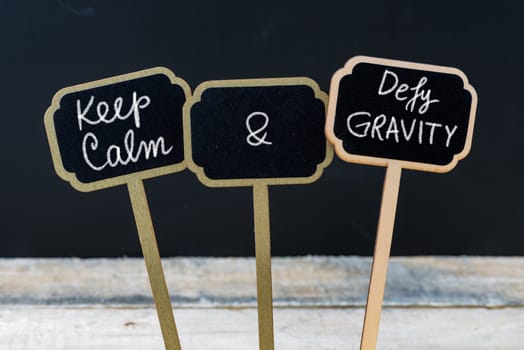 Keep Calm and Defy Gravity message written with chalk on mini blackboard labels, defocused chalkboard and wooden table in background. Fun and humor concept