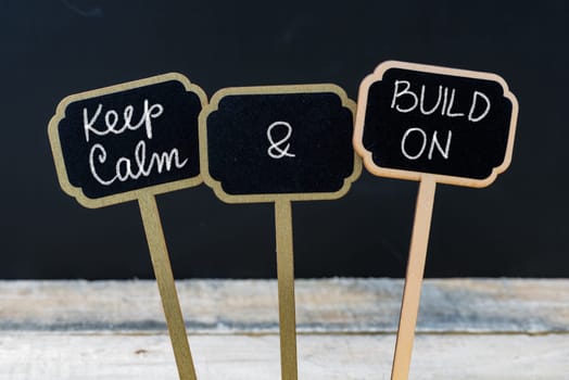 Keep Calm and Build On message written with chalk on mini blackboard labels, defocused chalkboard and wooden table in background. Fun and humor concept