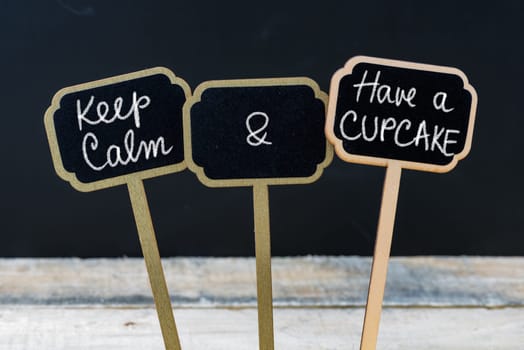 Keep Calm and Have A Cupcake message written with chalk on mini blackboard labels, defocused chalkboard and wooden table in background. Fun and humor concept