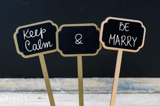 Keep Calm and Be Marry message written with chalk on mini blackboard labels, defocused chalkboard and wooden table in background. Fun and humor concept