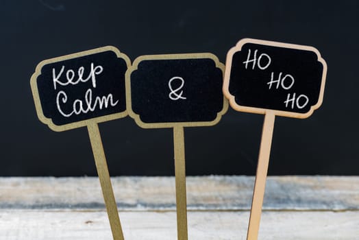 Keep Calm and HO HO HO message written with chalk on mini blackboard labels, defocused chalkboard and wooden table in background. Fun and humor concept