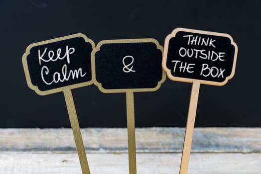 Keep Calm and Think Outside The Box message written with chalk on mini blackboard labels, defocused chalkboard and wooden table in background. Fun and humor concept