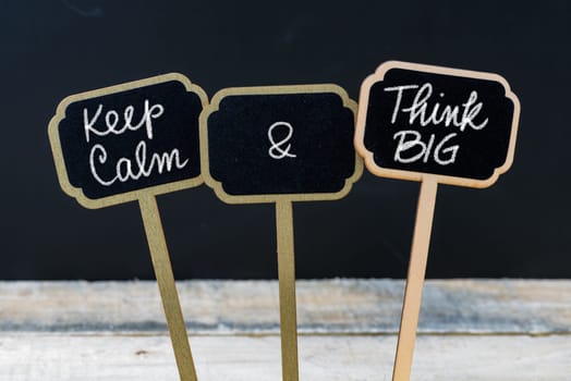 Keep Calm and Think Big message written with chalk on mini blackboard labels, defocused chalkboard and wooden table in background. Fun and humor concept