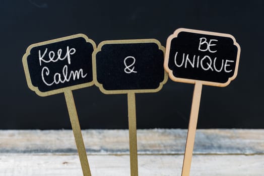 Keep Calm and Be Unique message written with chalk on mini blackboard labels, defocused chalkboard and wooden table in background. Fun and humor concept
