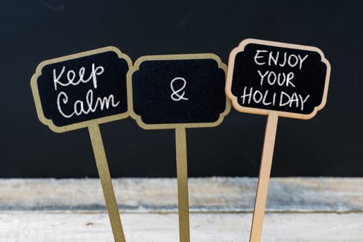 Keep Calm and Enjoy Your Holiday message written with chalk on mini blackboard labels, defocused chalkboard and wooden table in background. Fun and humor concept