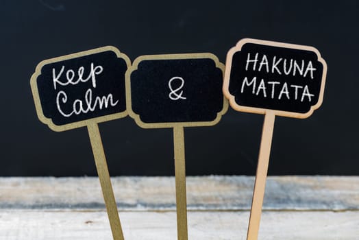 Keep Calm and Hakuna Matata message written with chalk on mini blackboard labels, defocused chalkboard and wooden table in background. Fun and humor concept