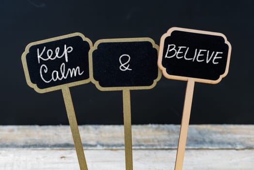 Keep Calm and Believe message written with chalk on mini blackboard labels, defocused chalkboard and wooden table in background. Fun and humor concept