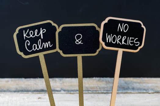 Keep Calm and No Worries message written with chalk on mini blackboard labels, defocused chalkboard and wooden table in background. Fun and humor concept